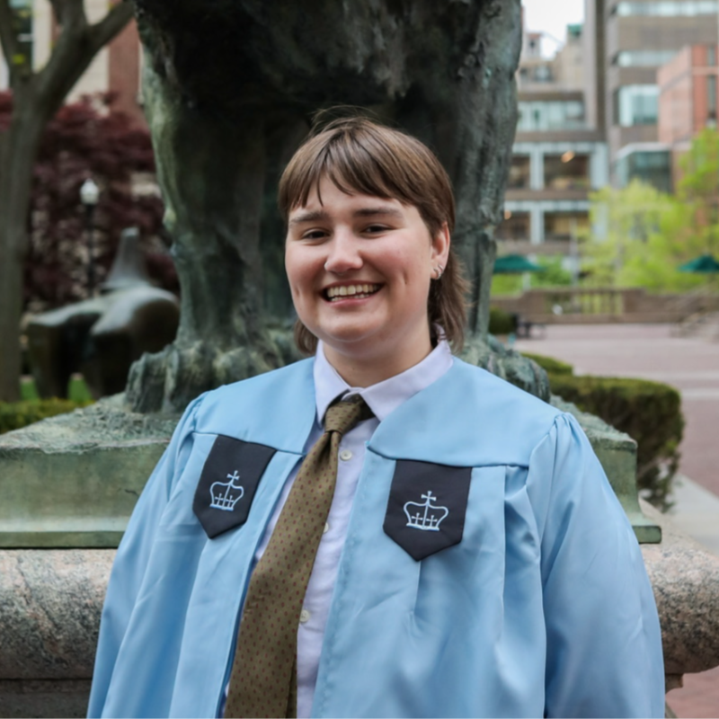 Headshot of LJ Mooney smiling in a light blue graduation stole, with a statue and campus greenery behind them.
